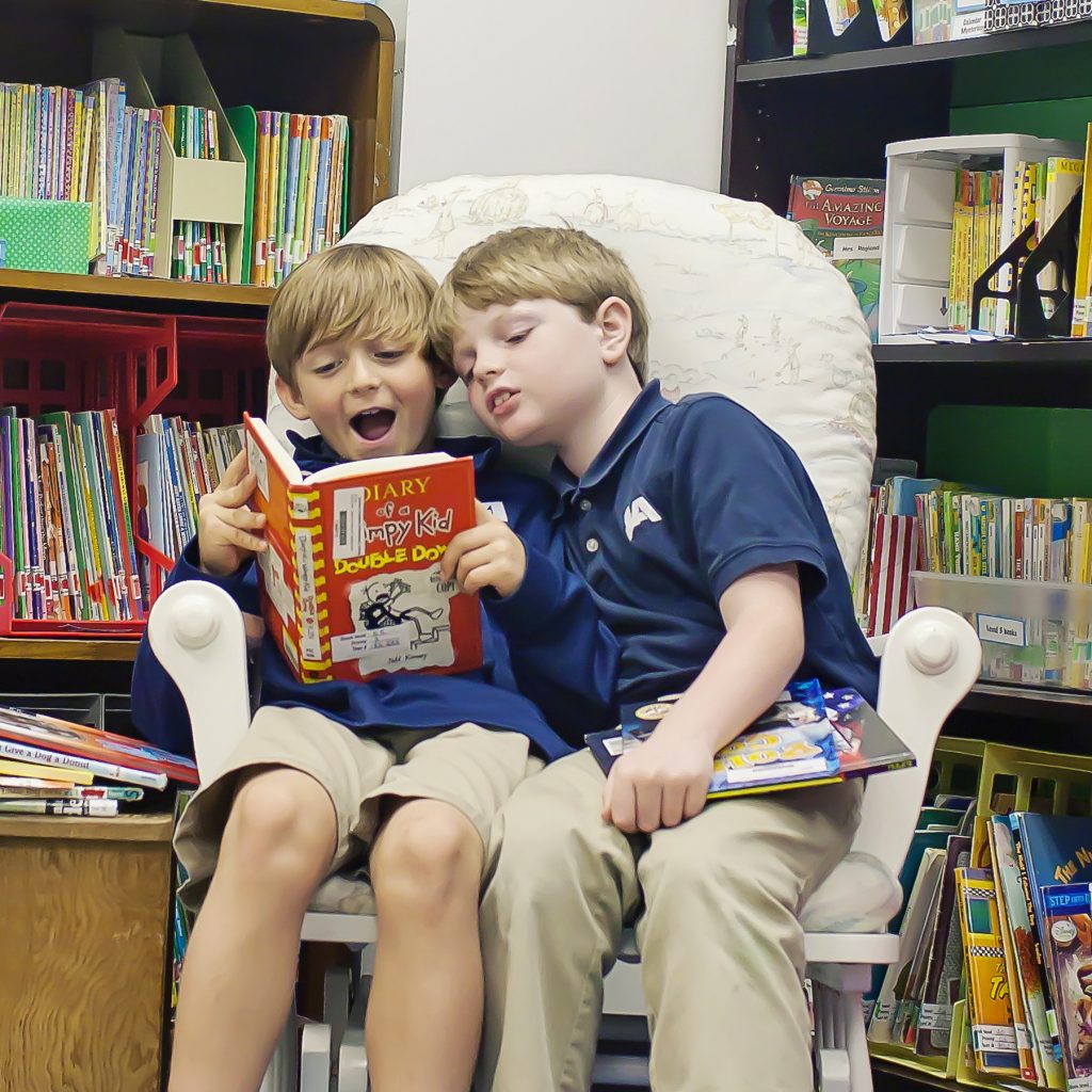 Two elementary age boys read a book together while sitting in a comfortable chair in the Lower School library.